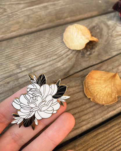Floral pin in a beautiful peony tattoo style design, held by a hand on a wooden surface with dried leaves.