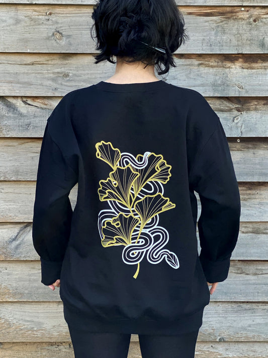 Lu Loram-Martin is wearing a black sweatshirt with a gold and white illustration of a snake amongst gingko leaves 